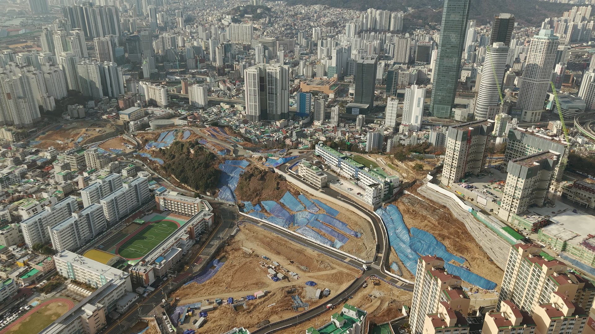 파란 덮개가 덮인 언덕의 빈터와 그 곁의 학교 운동장, 멀리 국제금융센터와 아파트들을 드론으로 내려다본 모습